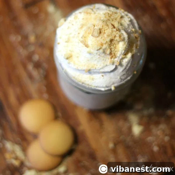Banana Cream Pie Smoothie Ready to Serve A glass of Banana Cream Pie Smoothie garnished with whipped cream and a vanilla wafer on a rustic wooden background.