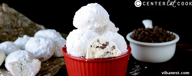chocolate_chip_snowball_cookies1.jpg A small pile of perfectly round, white powdered sugar-dusted chocolate chip snowball cookies in a festive setting.