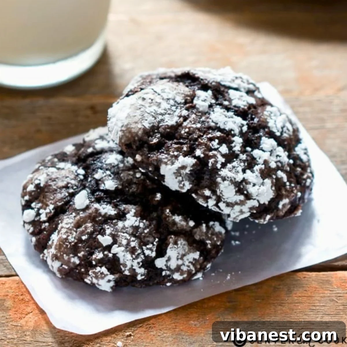 Velvet Chocolate Crinkle Cookies 2 Stack of four cakey chocolate crinkle cookies, heavily coated in powdered sugar, resting on a white plate with a subtle gray background. The cookies show beautiful white cracks contrasting with their dark chocolate interior.