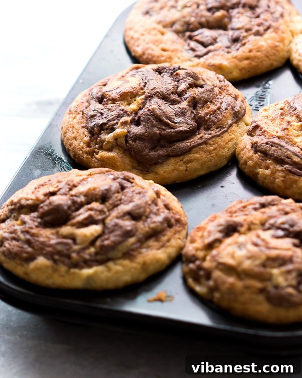 nutella-swirled-banana-muffins-2.jpg Close-up of baked Nutella Swirled Banana Muffins cooling on a wire rack, showcasing golden tops and rich Nutella patterns.