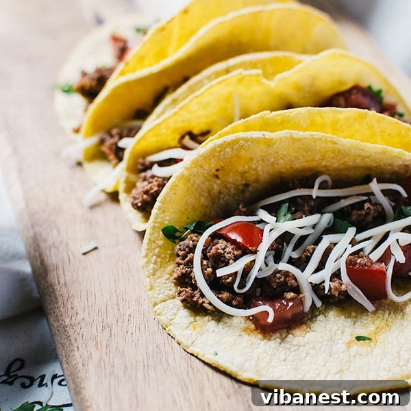 A bowl of slow cooker beef taco meat, accompanied by a selection of fresh toppings like cheese, tomatoes, and cilantro, with tortillas in the background