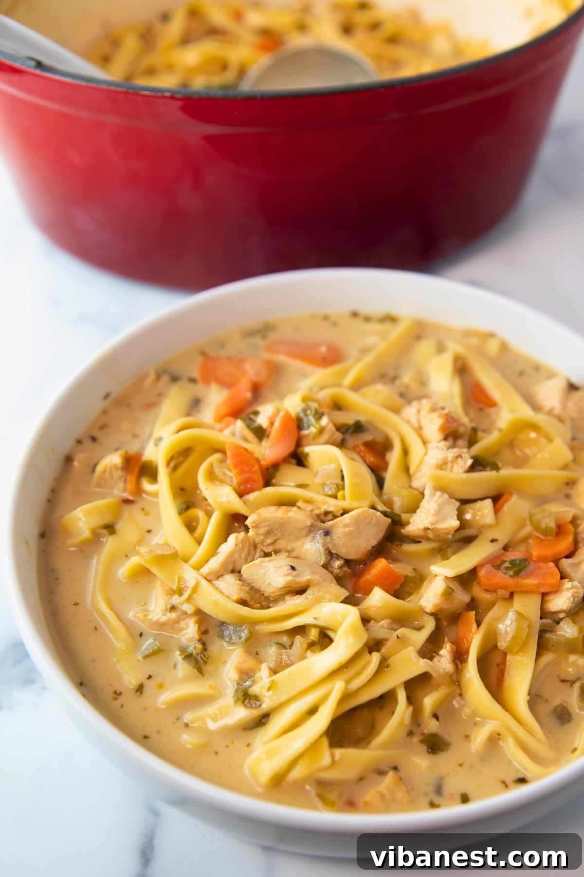 Comforting Creamy Chicken Noodle Soup 2 A beautiful bowl of creamy chicken noodle soup, with a rustic pot visible in the soft-focus background, evoking warmth and comfort.
