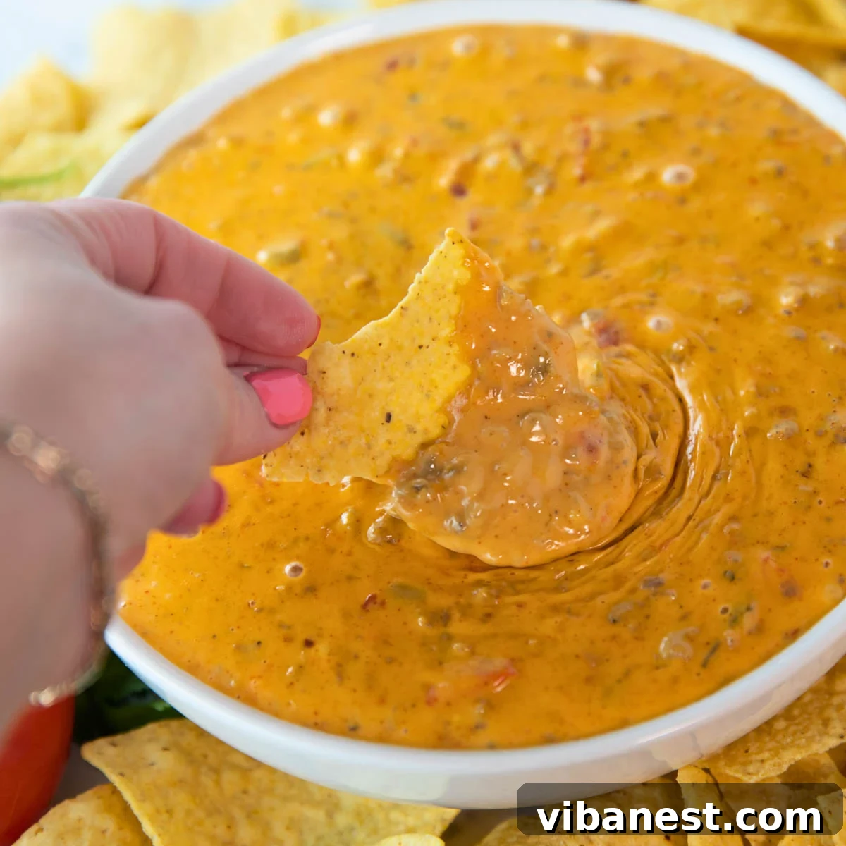 Crowd-Pleasing Rotel Sausage Dip 2 Woman's hand holding a chip and scooping creamy Easy Rotel Dip with Sausage from a bowl, surrounded by tortilla chips, ready for a party.
