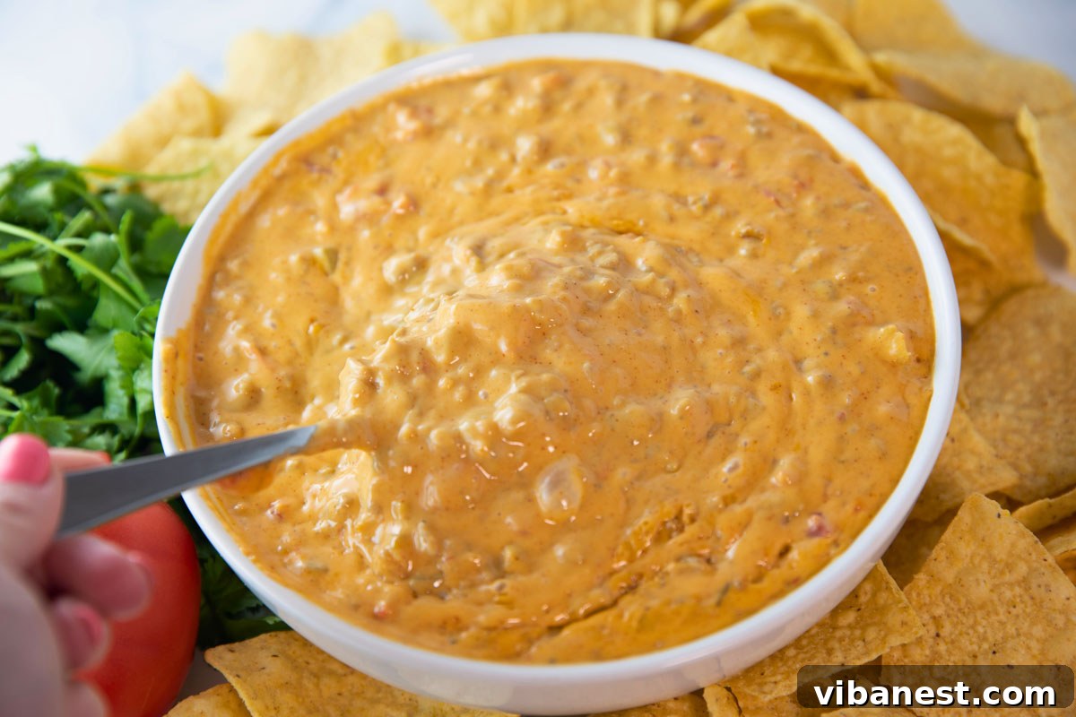 Crowd-Pleasing Rotel Sausage Dip 7 Woman's hand holding a spoon to stir creamy easy Rotel dip with sausage in a bowl, garnished with fresh cilantro and surrounded by tortilla chips.