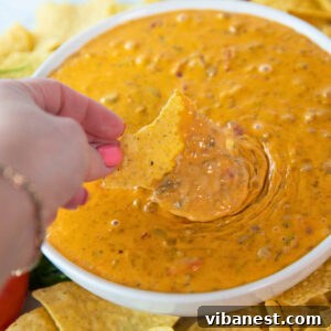 Close-up of a woman's hand holding a chip and scooping creamy Easy Rotel Dip with Sausage, with a bowl surrounded by tortilla chips.