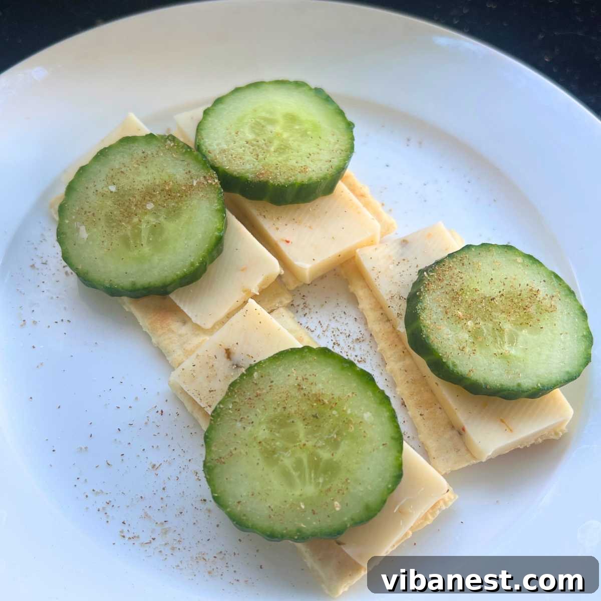 The Friday Five May 31 2024 4 A close-up of a cracker topped with a slice of cheese, cucumber, and a sprinkle of spices, showcasing a fresh and appealing snack.