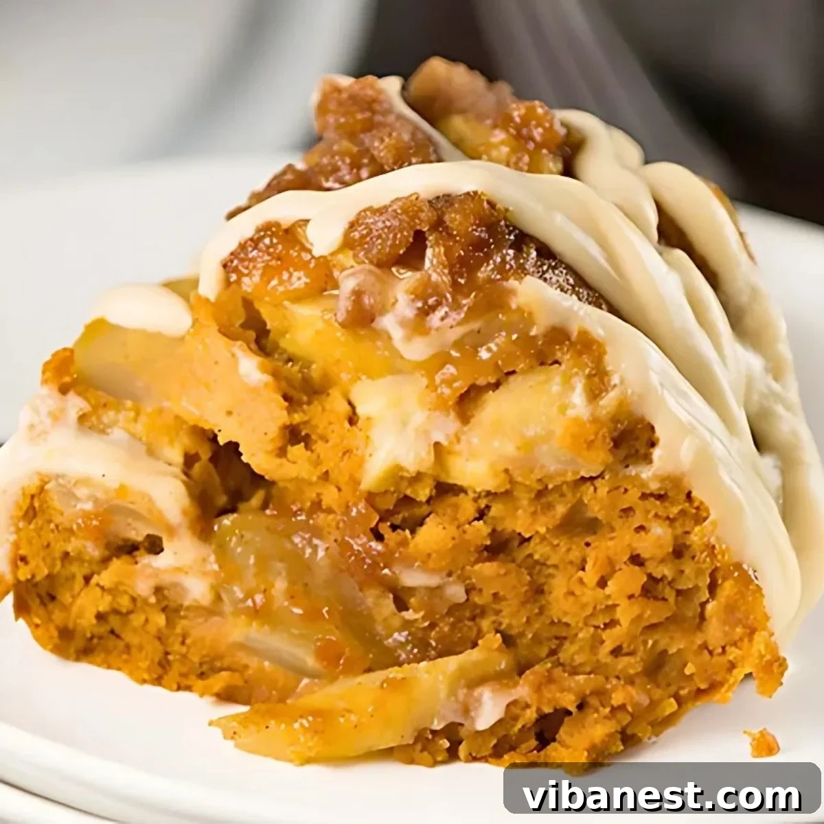 Harvest Spice Pumpkin Apple Cake 2 A stunning pumpkin apple spice Bundt cake adorned with a generous drizzle of cream cheese glaze, resting elegantly on a pristine white plate, surrounded by autumnal décor.