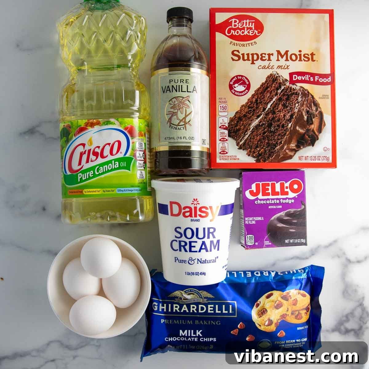 Decadent Double Chocolate Bundt Cake 5 A vibrant flat lay showcasing all the ingredients required for the rich chocolate chocolate chip bundt cake: a box of Devil's Food cake mix, instant chocolate pudding, eggs, vanilla, sour cream, vegetable oil, water, and milk chocolate chips.