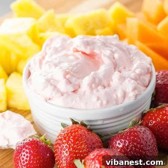 A beautifully presented bowl of creamy fruit dip, garnished with a fresh mint sprig, surrounded by a vibrant and colorful assortment of fresh fruits like strawberries, blueberries, grapes, and pineapple chunks on a white platter