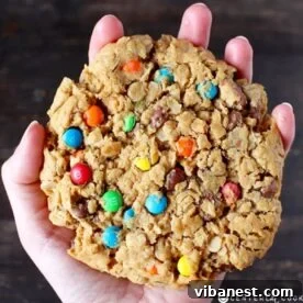A person's hand proudly holding a colossal, thick giant monster cookie, showcasing its many colorful mix-ins.
